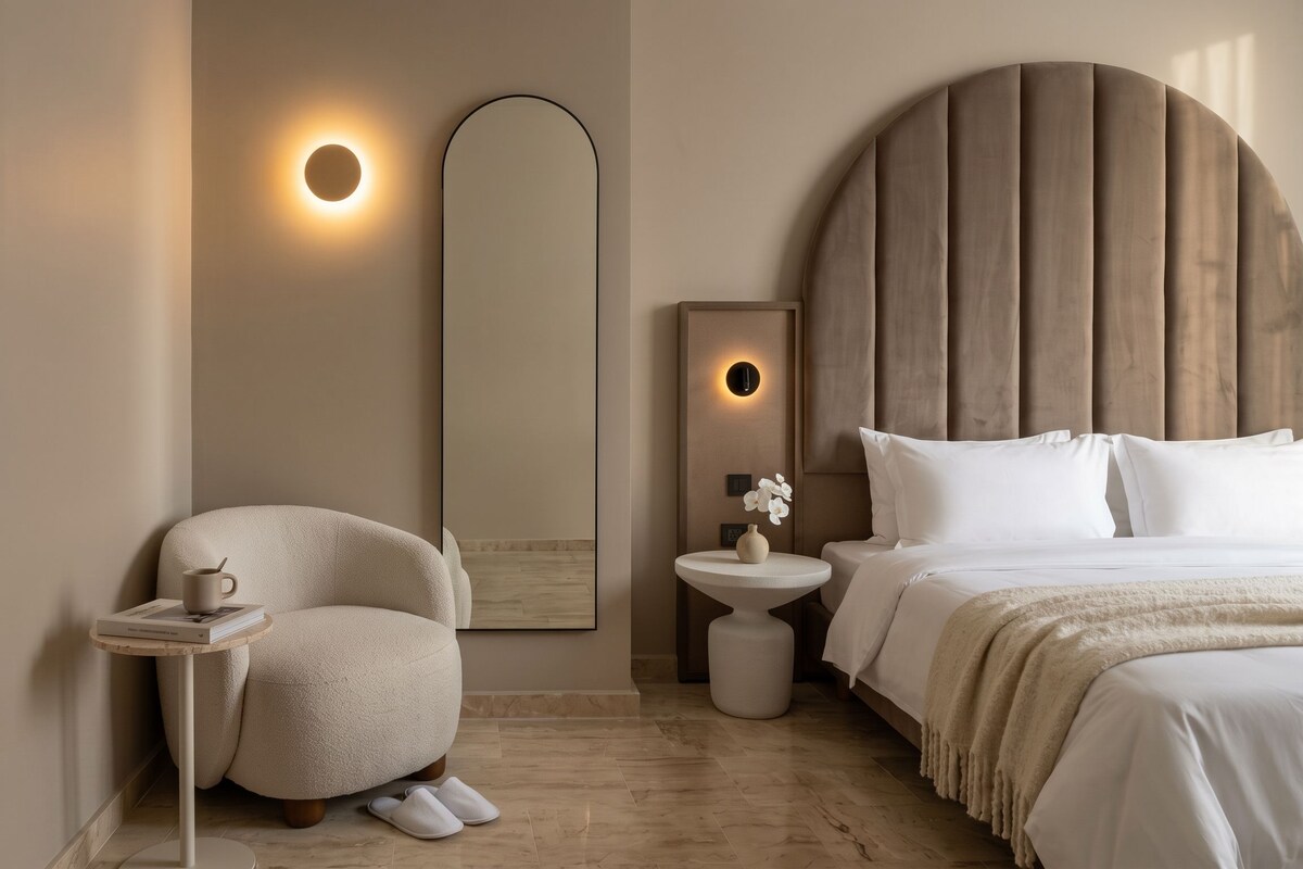 A modern bedroom with a large bed, a round side table, a chair, and a wall-mounted mirror.