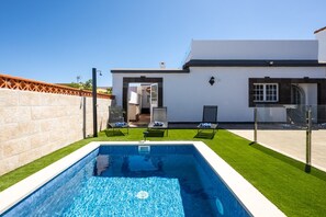 A modern house with a swimming pool, two lounge chairs, and a clear sky.