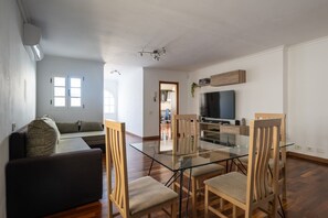 A modern living room with a glass dining table, wooden chairs, a sofa, a flat-screen TV, and a ceiling fan.