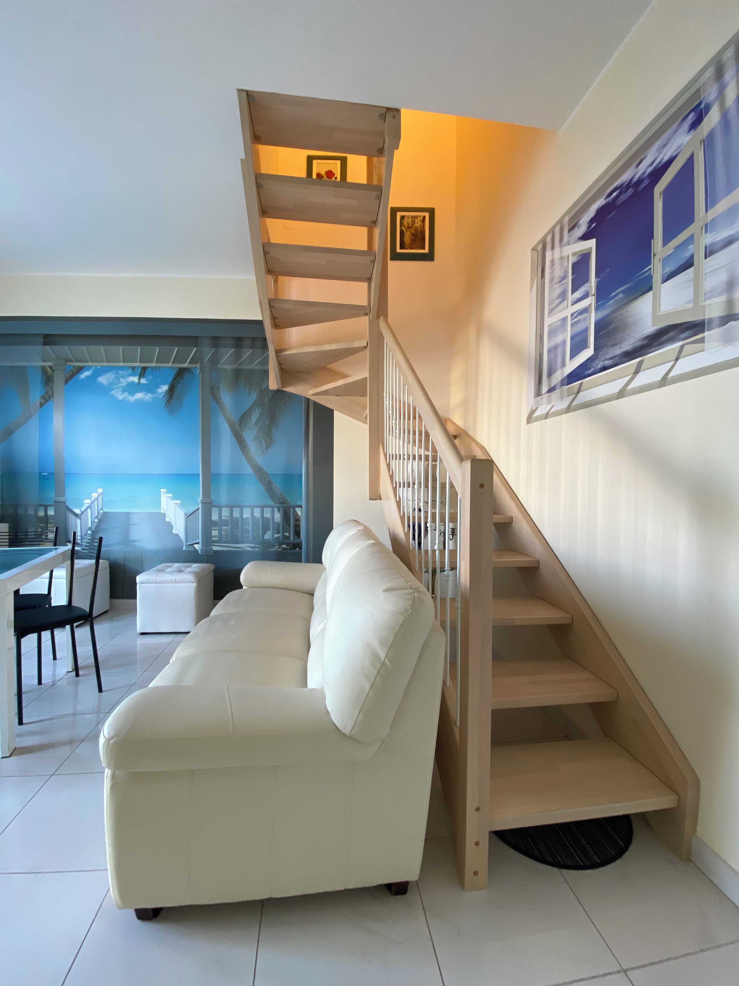 A modern living room with a white sofa, a wooden staircase, and a view of the ocean through a glass door.