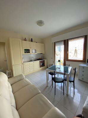 A living room with a beige sofa, a dining table with chairs, a kitchen with cabinets and appliances, and a window with curtains. - Fairmont Residence Elisabetta by Ca-sa Check (Chioggia)