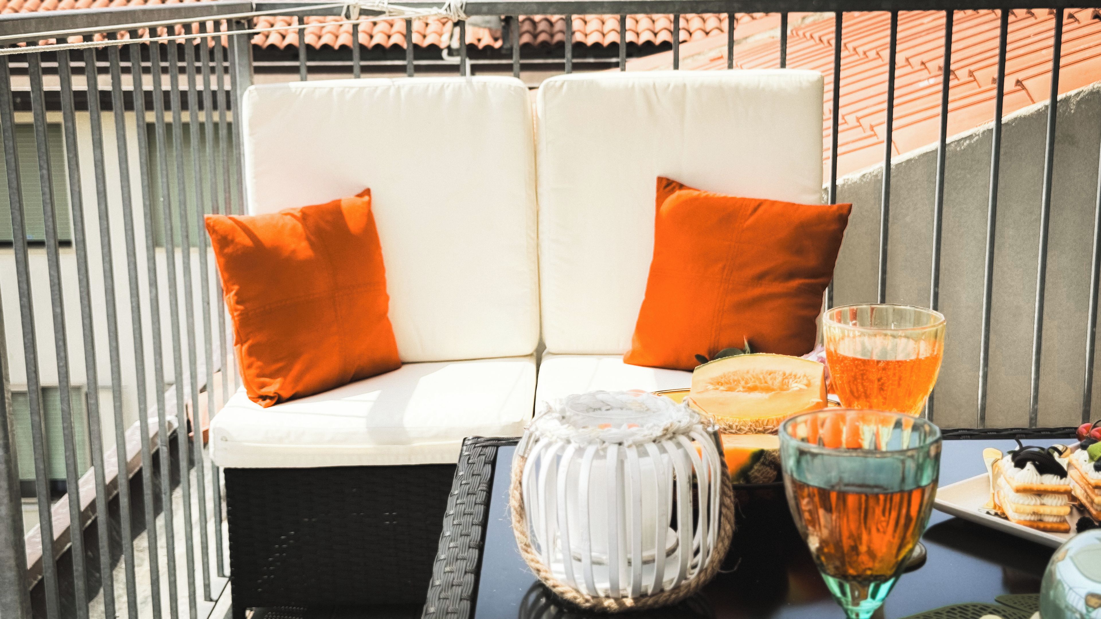 Een balkon met een witte bank, oranje kussens, een glazen tafel met een glas amberkleurig vocht, een witte lantaarn, en een bord met eten.