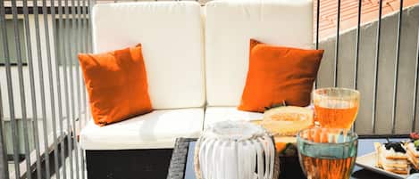 Een balkon met een witte bank, oranje kussens, een glazen tafel met een glas amberkleurig vocht, een witte lantaarn, en een bord met eten.