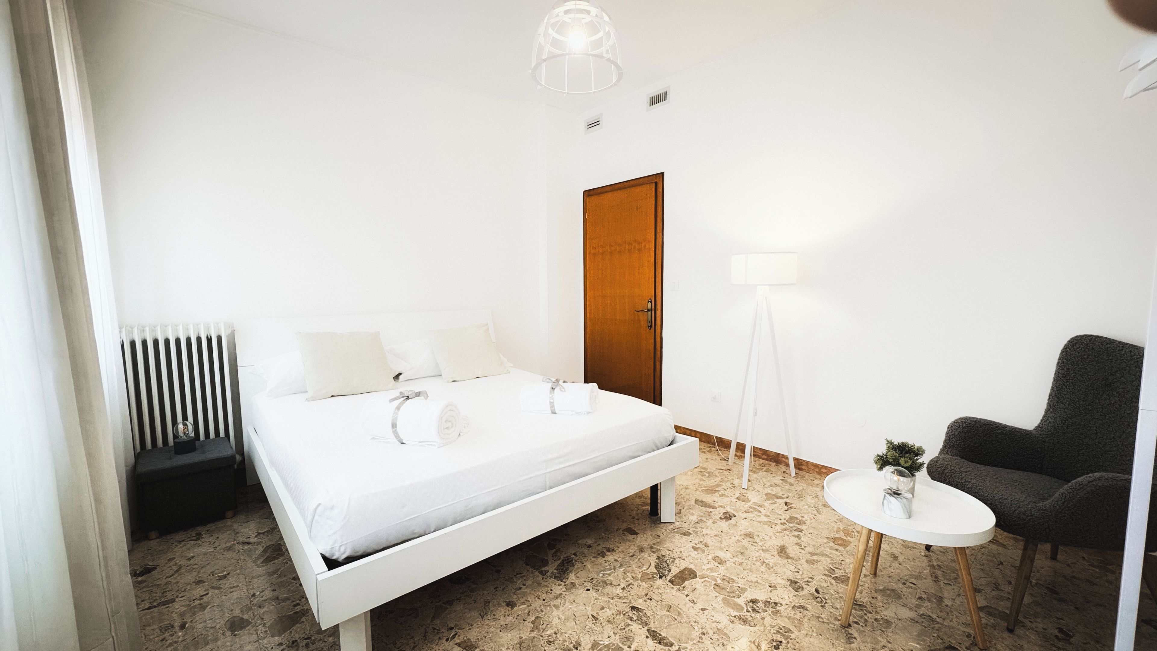 Un dormitorio con cama, mesita de noche, una silla, lámpara de pie y radiador.