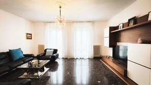Un salon moderne avec un canapé en cuir noir, une table basse en verre, un meuble TV en bois et un lustre.