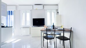 A modern kitchen with a dining area, white cabinetry, a flat-screen TV, and a window with blue curtains. - Farmont Residence Alice by Ca-sa Check (Chioggia)