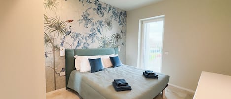 A bedroom with a bed, pillows, a nightstand, a wall-mounted light, and a door leading to a balcony.