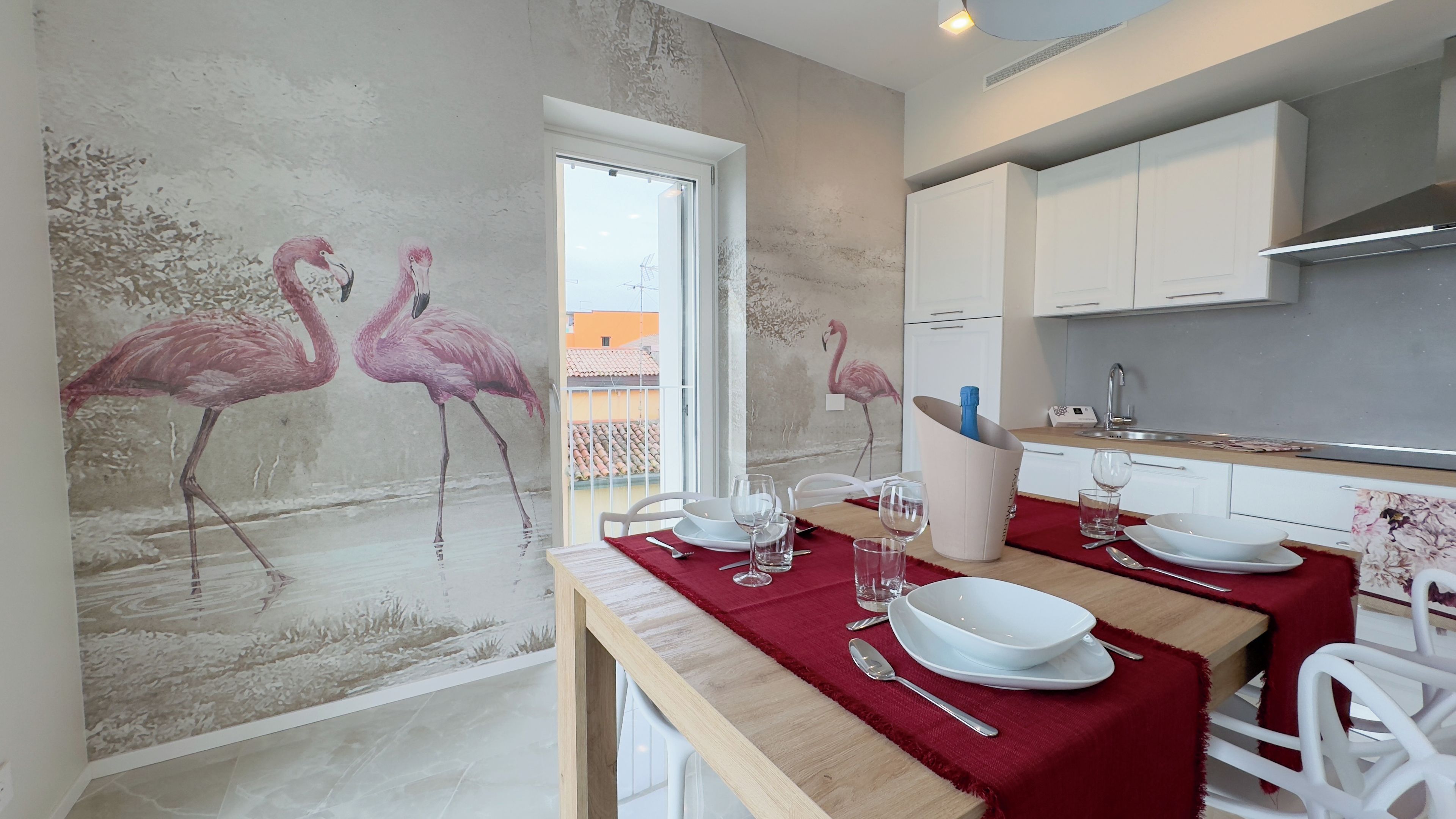 Uma área de refeições com mesa posta para dois, cozinha com armários brancos e um mural de flamingos na parede.