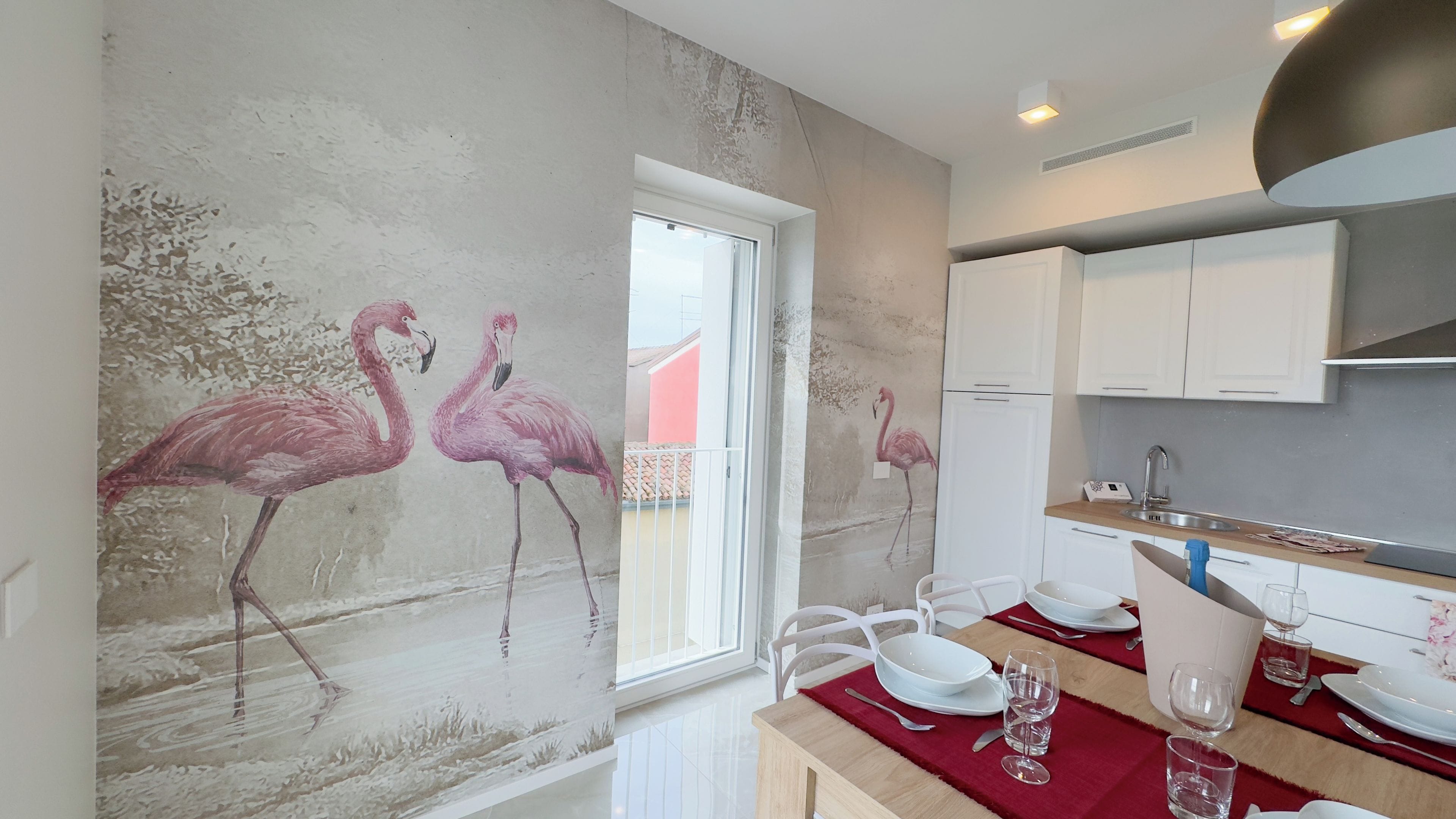 Cozinha moderna com armários brancos, área de jantar com toalha vermelha e um mural na parede com flamingos.