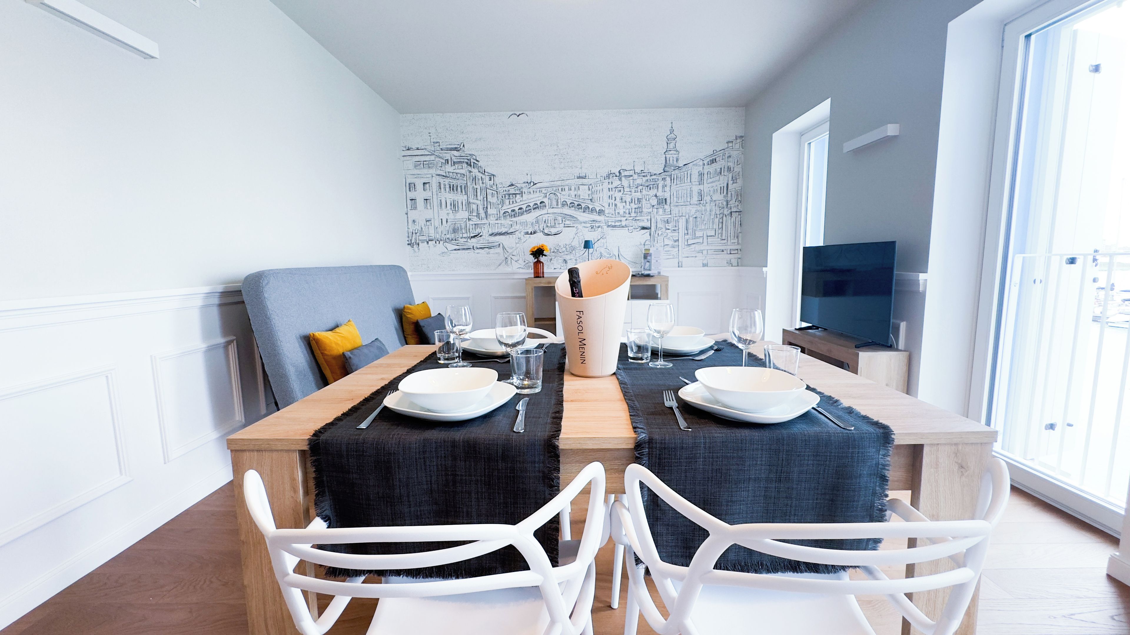 Un coin repas avec une table dressée pour deux personnes, une fresque représentant un paysage urbain sur le mur et un téléviseur posé sur un meuble.