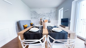 Un área de comedor con una mesa preparada para dos, un mural con vista a la ciudad en la pared y un televisor sobre un soporte.