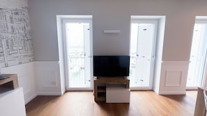 Una habitación con piso de madera, un televisor de pantalla plana sobre un pedestal y dos puertas que dan a un balcón con vistas a barcos.