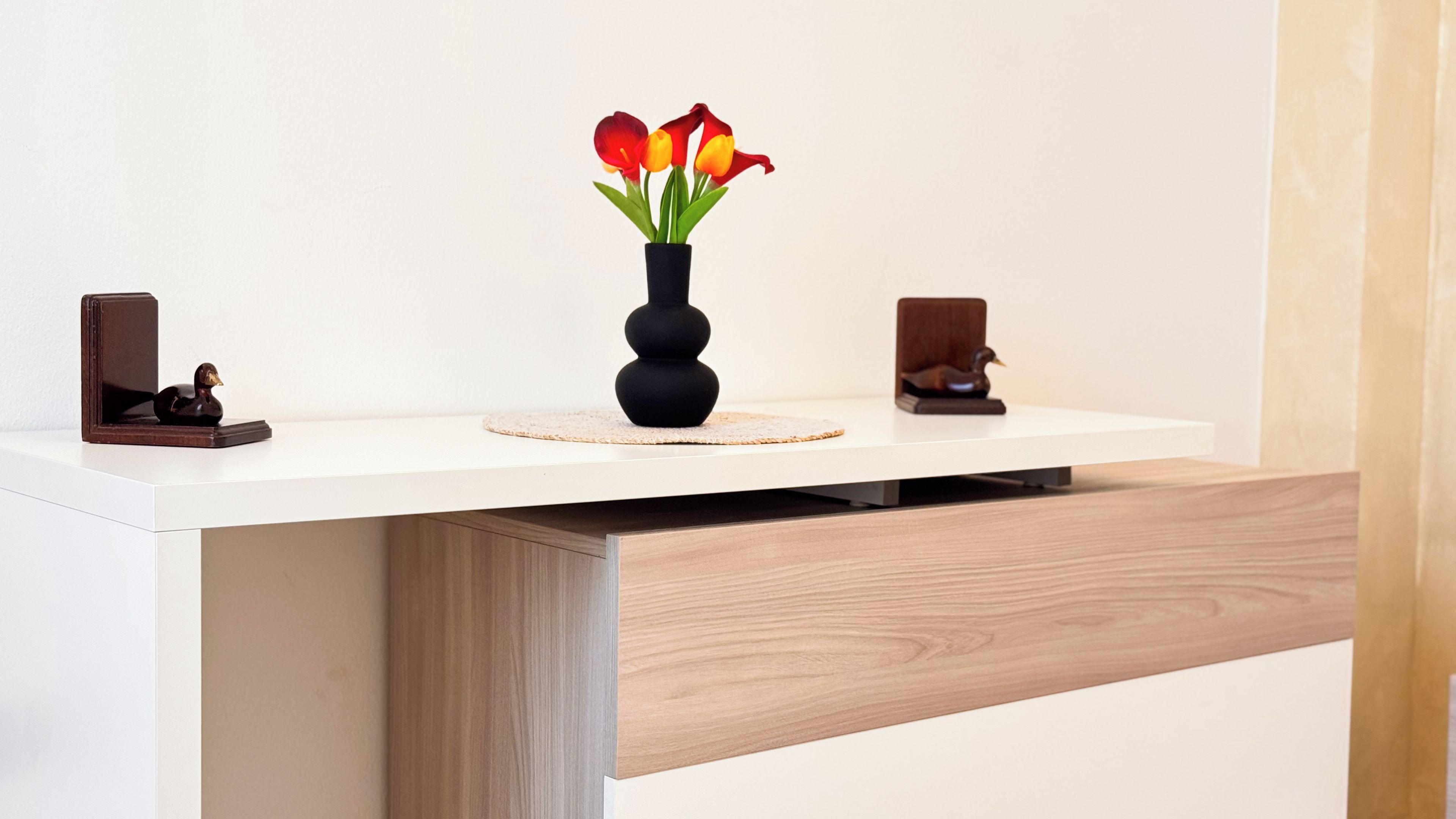 Uma escrivaninha moderna com um vaso de flores, uma figurinha de pato e um pato de madeira sobre uma superfície branca.