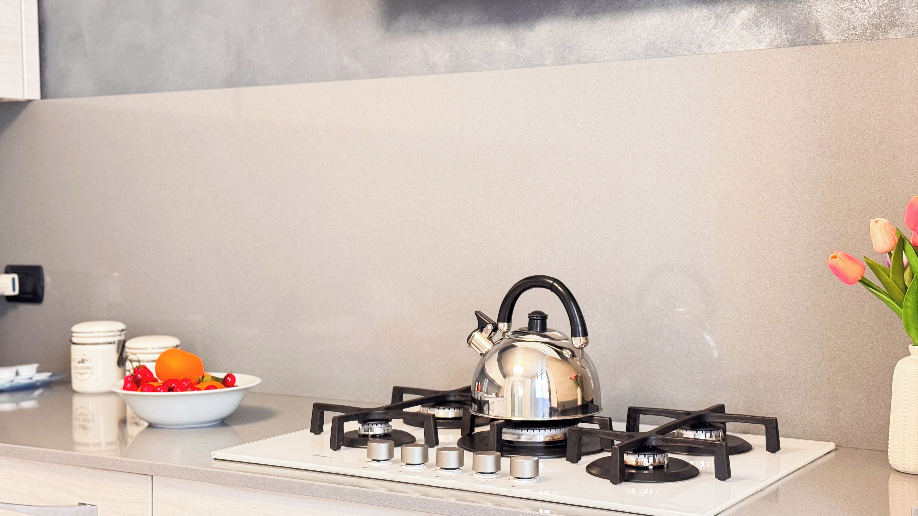 Una cocina con estufa a gas, hervidor, un cuenco de fruta y un jarrón con flores.
