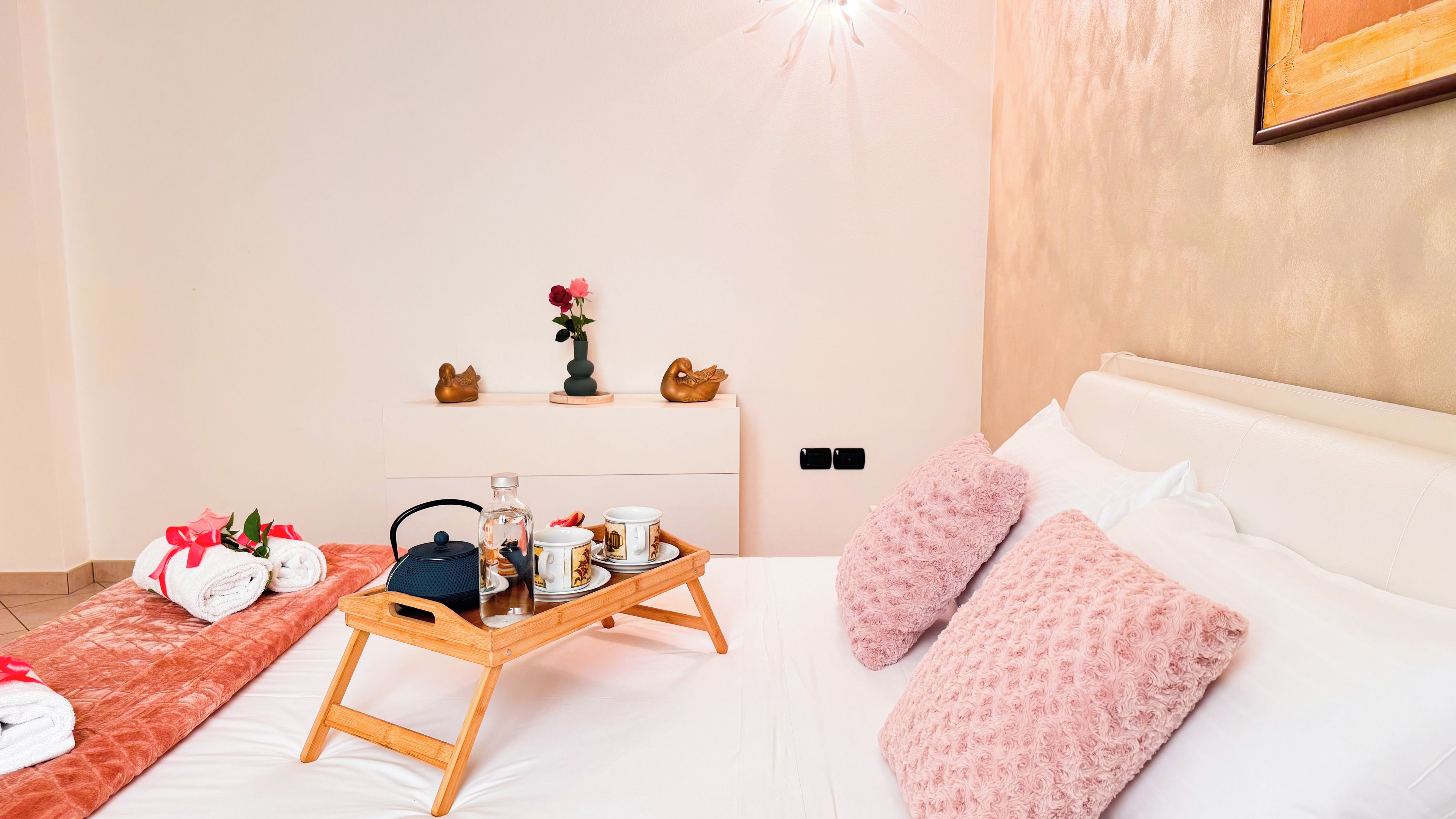Un dormitorio ordenado con cama, una mesita de noche con una bandeja de desayuno, una silla y un pequeño estante con adornos.