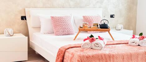Una cama bien tendida con ropa de cama rosa y blanca, una bandeja con té y aperitivos, y toallas enrolladas con motivos de rosas sobre una mesita de noche.
