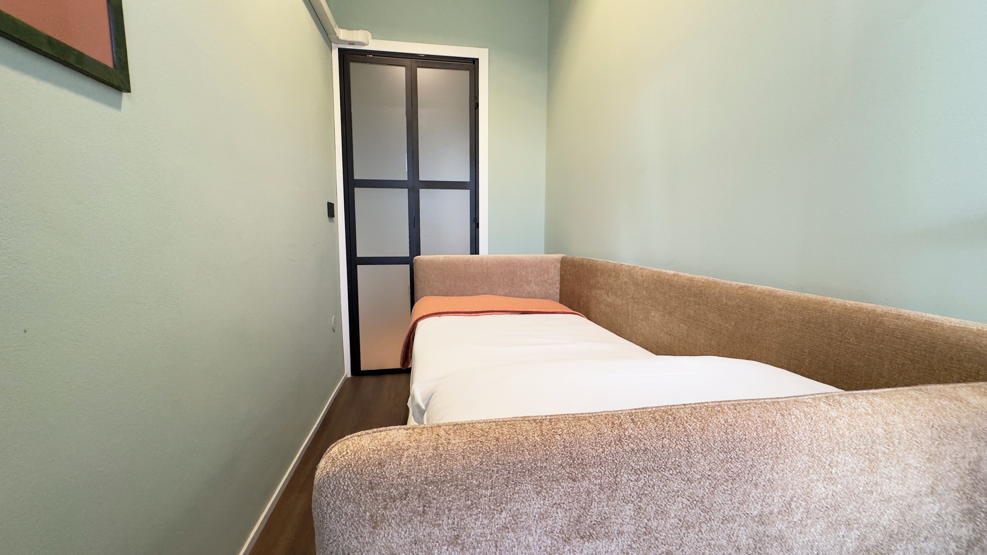 A narrow hallway with a bed at the end, a door with frosted glass panels, and a light-colored wall.