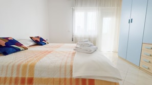 Quarto com cama, almofadas, pilha de toalhas dobradas, guarda-roupa e janela com cortinas.