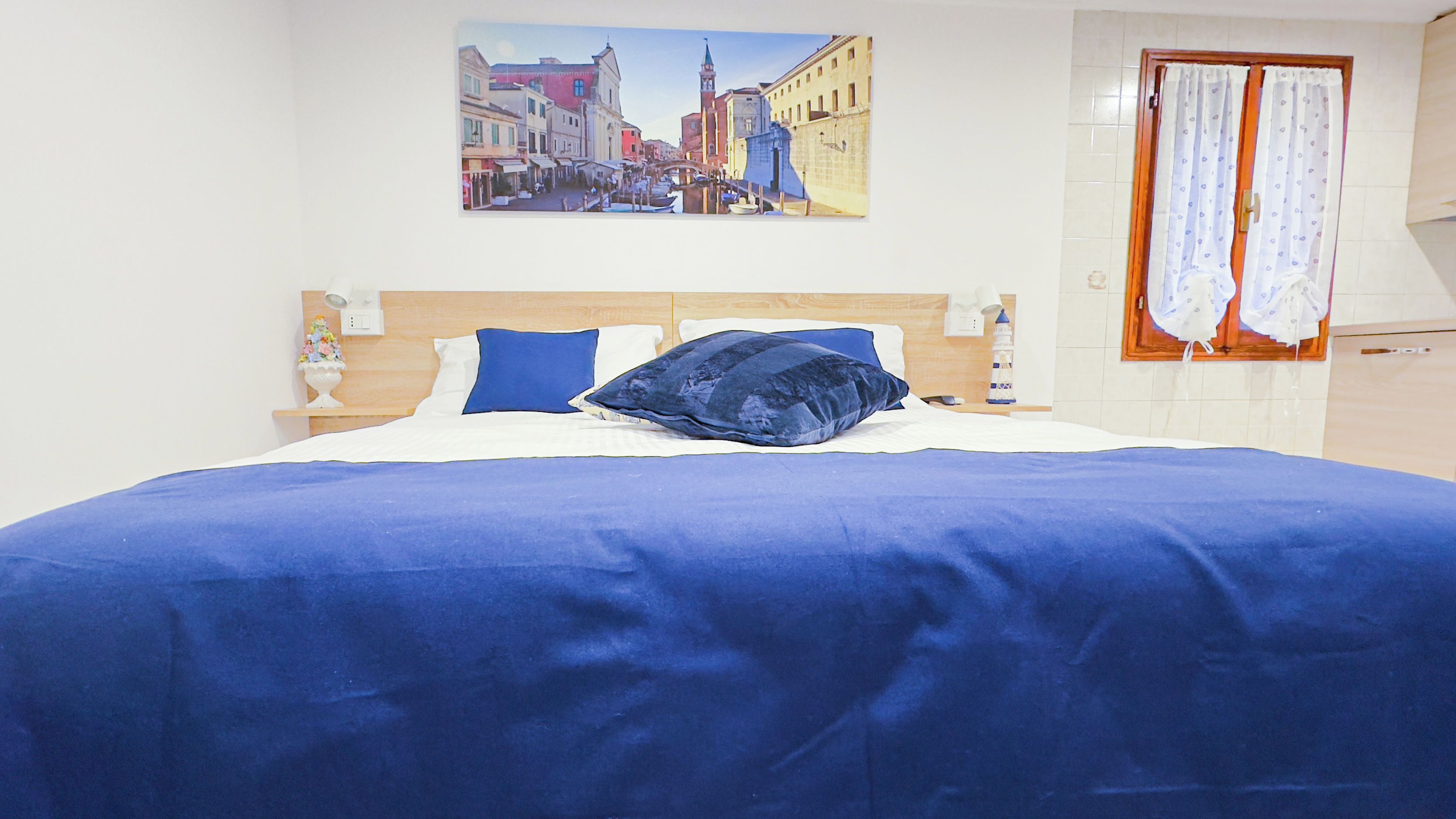 Un dormitorio con cama, mesita de noche, lámpara, cuadro de un paisaje urbano y ventana con cortinas.