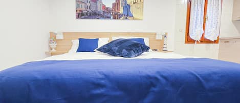 Une chambre à coucher avec un lit, une table de chevet, une lampe, un tableau représentant un paysage urbain et une fenêtre avec des rideaux.