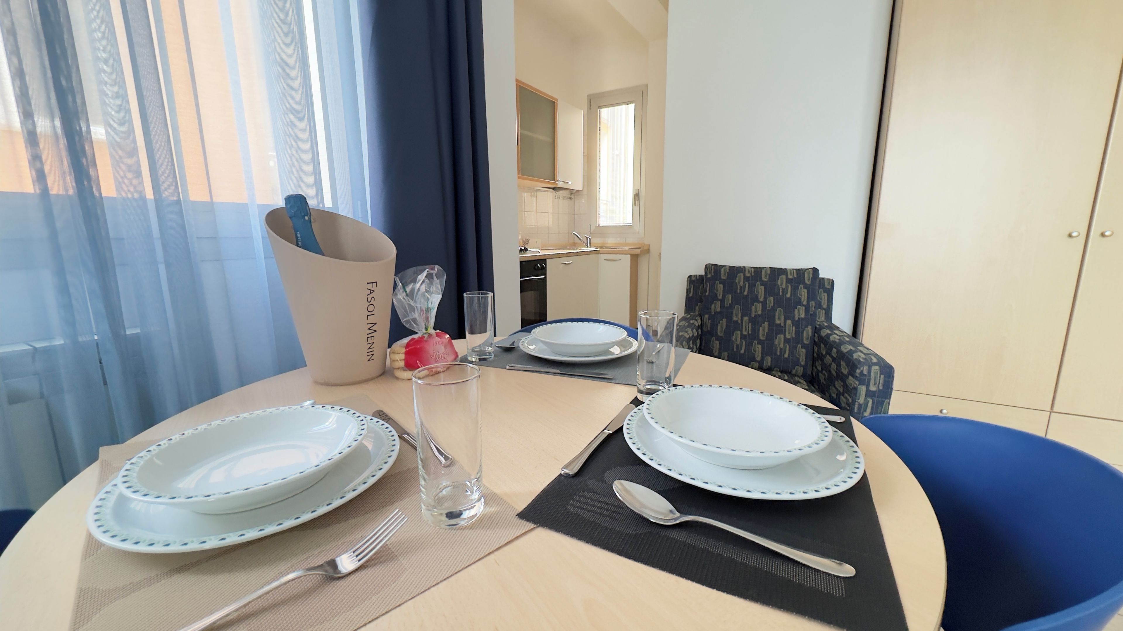Mesa de jantar posta para dois, com pratos, talheres e copos, um vaso com flores e um saco de lanches.