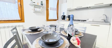 Un comedor con una mesa dispuesta para cuatro personas, incluyendo platos, tazones, vasos y cubiertos. Hay sillas alrededor de la mesa, y se ve una zona de cocina al fondo.