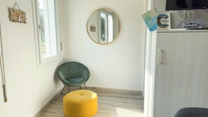 Una habitación con piso de madera, una silla verde, un reposapiés amarillo, un espejo y un mueble con televisor.