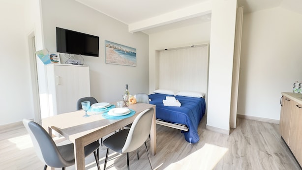 A compact hotel room with a bed, a dining area with a table and chairs, a flat-screen TV mounted on the wall, and a map on the cabinet. - Onda Blu House by Ca-sa Check (Chioggia)