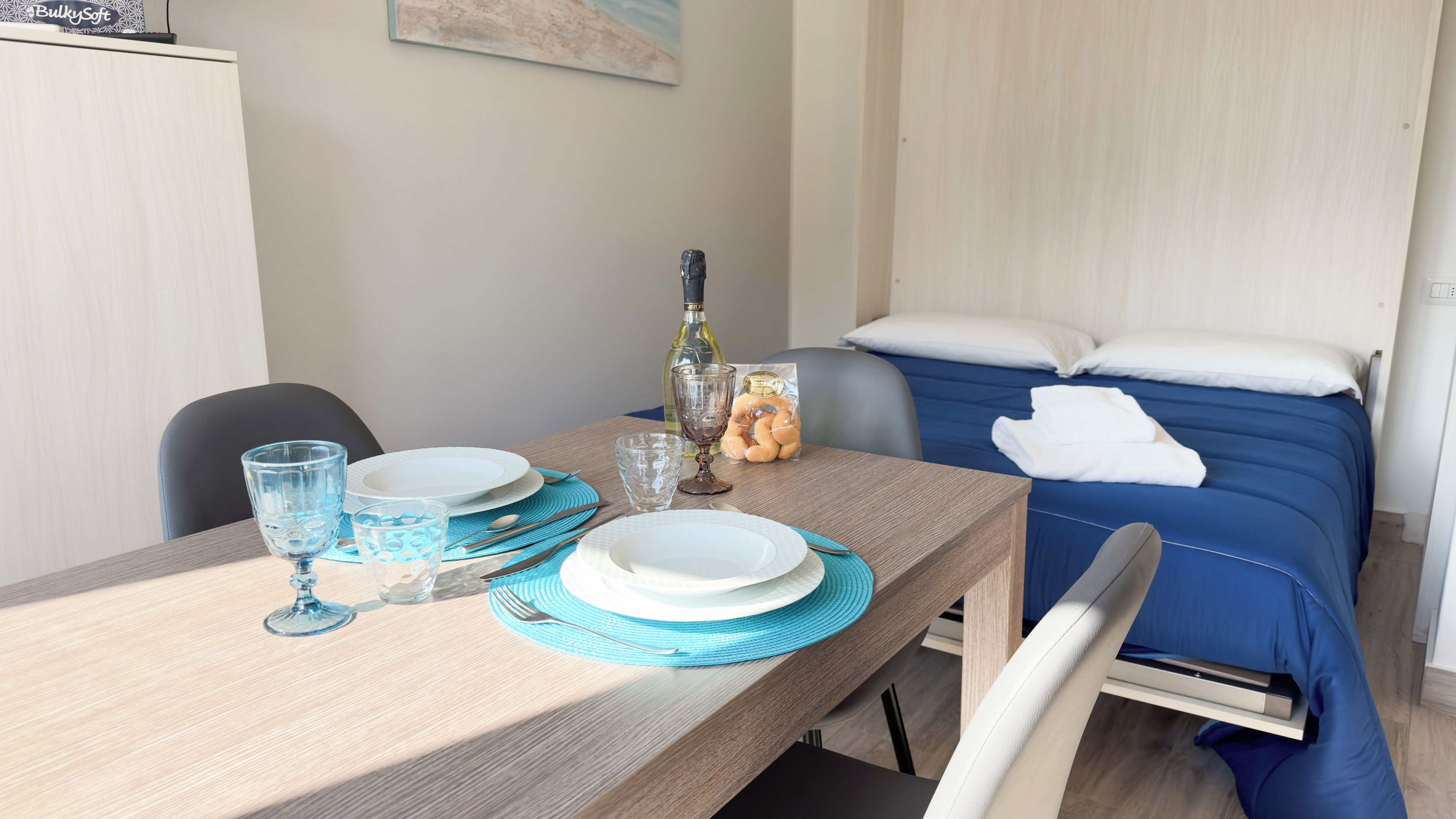 Une table à manger bien dressée, avec des assiettes, des verres et une bouteille, un lit avec une literie bleue et un tableau accroché au mur.