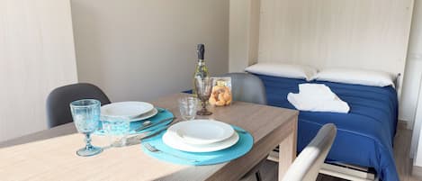 A neatly arranged dining table with plates, glasses, and a bottle, a bed with blue bedding, and a wall-mounted picture.