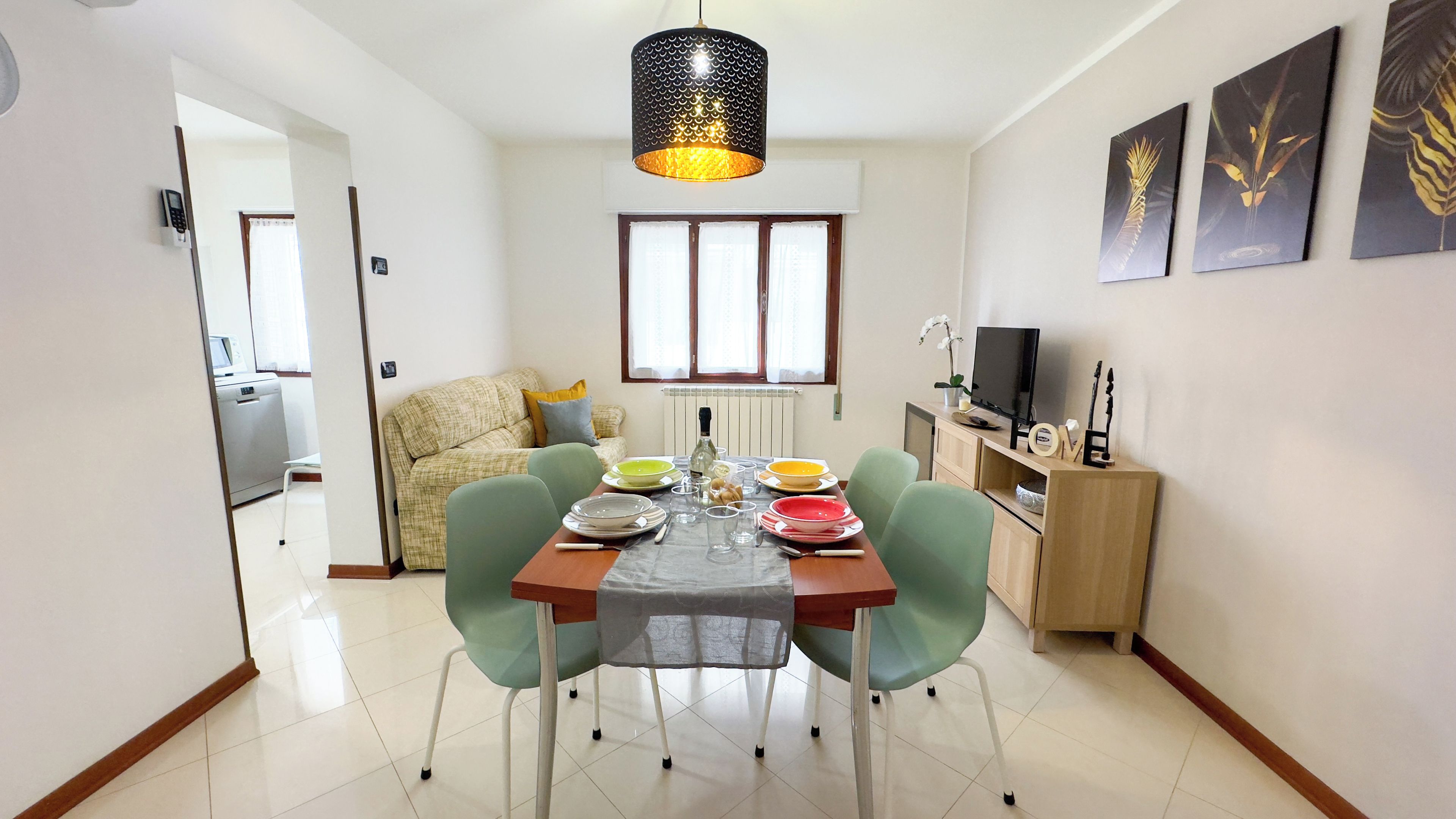 Een eetgedeelte met een tafel voor vier personen, stoelen, een hanglamp, een raam met gordijnen, een bank, een televisie en een kast met decoratieve objecten.
