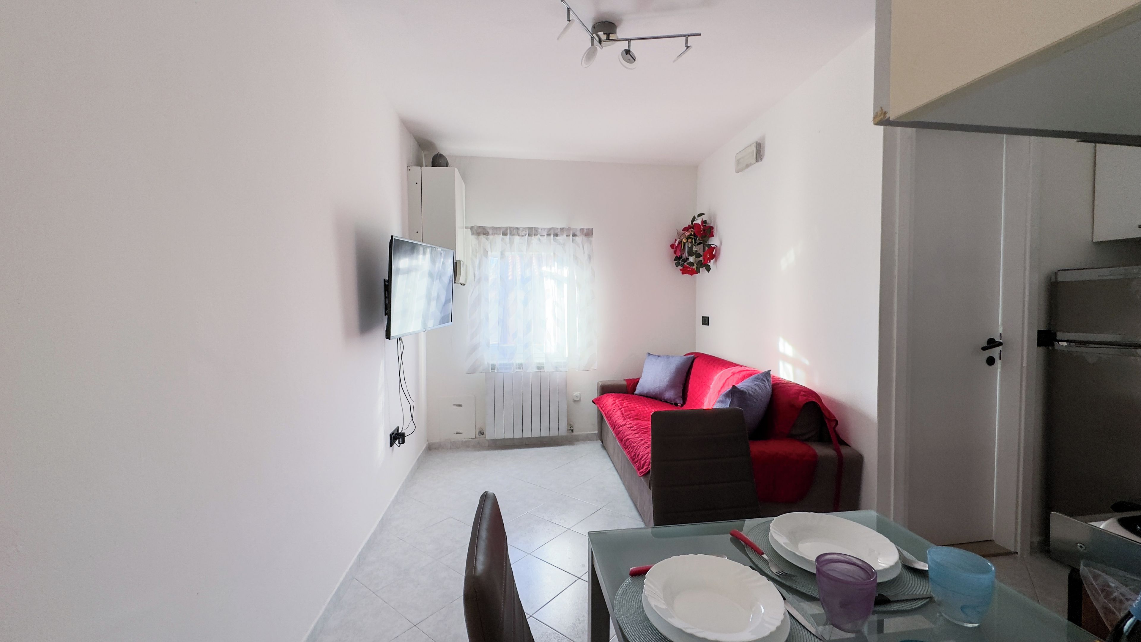 A compact living space with a dining table, a sofa with cushions, a television mounted on the wall, and a refrigerator.