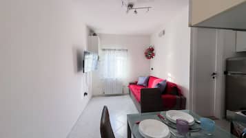 A compact living space with a dining table, a sofa with cushions, a television mounted on the wall, and a refrigerator.