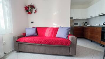 A modern living room with a red sofa, two pillows, a kitchen area with cabinets and appliances, and a decorative wall piece.