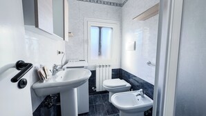Ein Badezimmer mit einem weißen Waschbecken, einer Toilette und einem Bidet. Über der Toilette befindet sich ein Fenster.