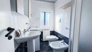 Casa de banho com lavatório branco, sanita e bidé. Existe uma janela sobre a sanita.