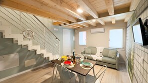 Uma área de jantar com mesa posta para quatro pessoas, escada com detalhe de roda decorativa e sofá em um espaço com vigas de madeira e janela.