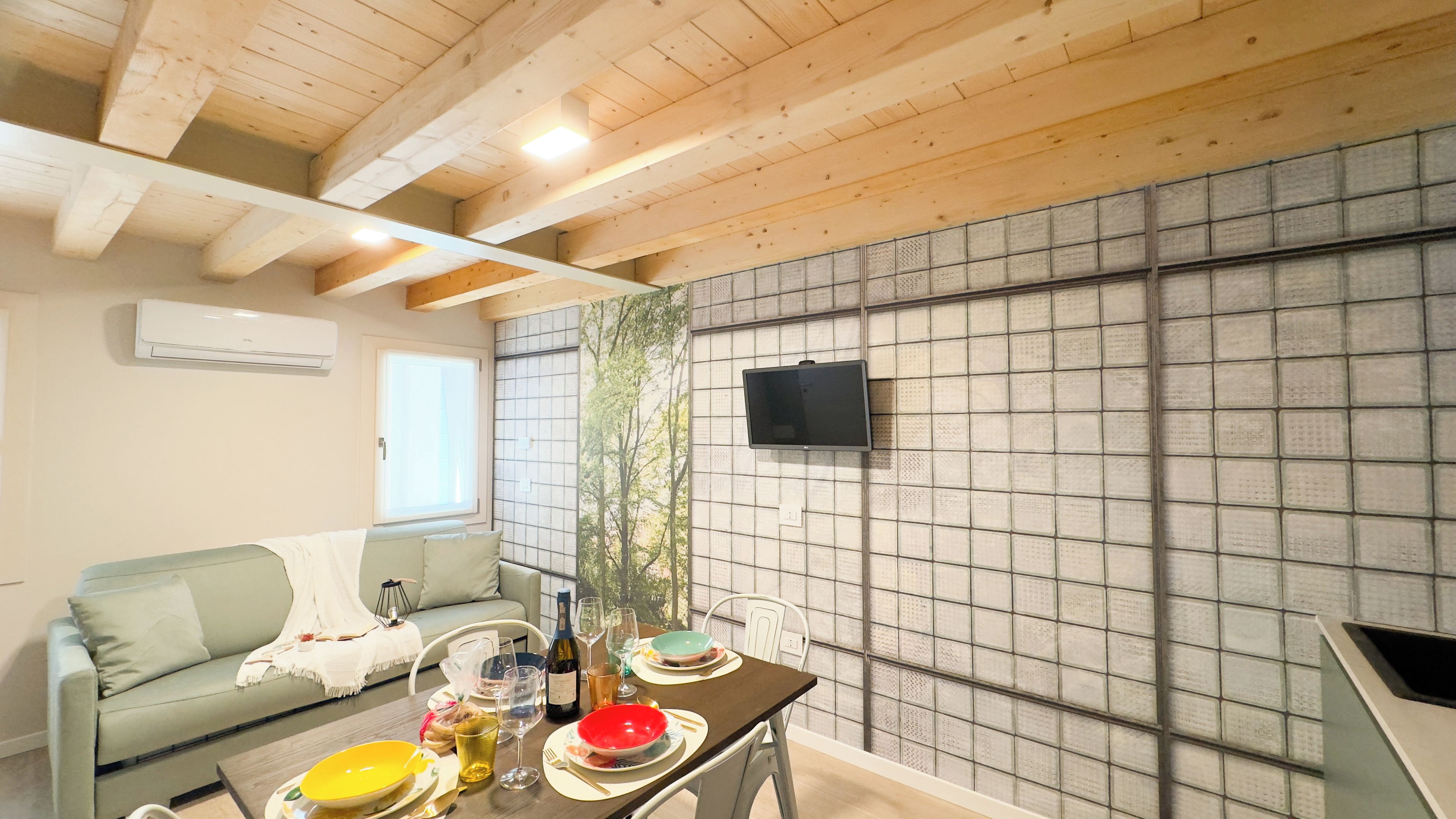 Quarto com teto de madeira, TV fixada em parede revestida de azulejos, mesa de jantar posta com pratos, copos e uma garrafa, e um sofá com almofada.