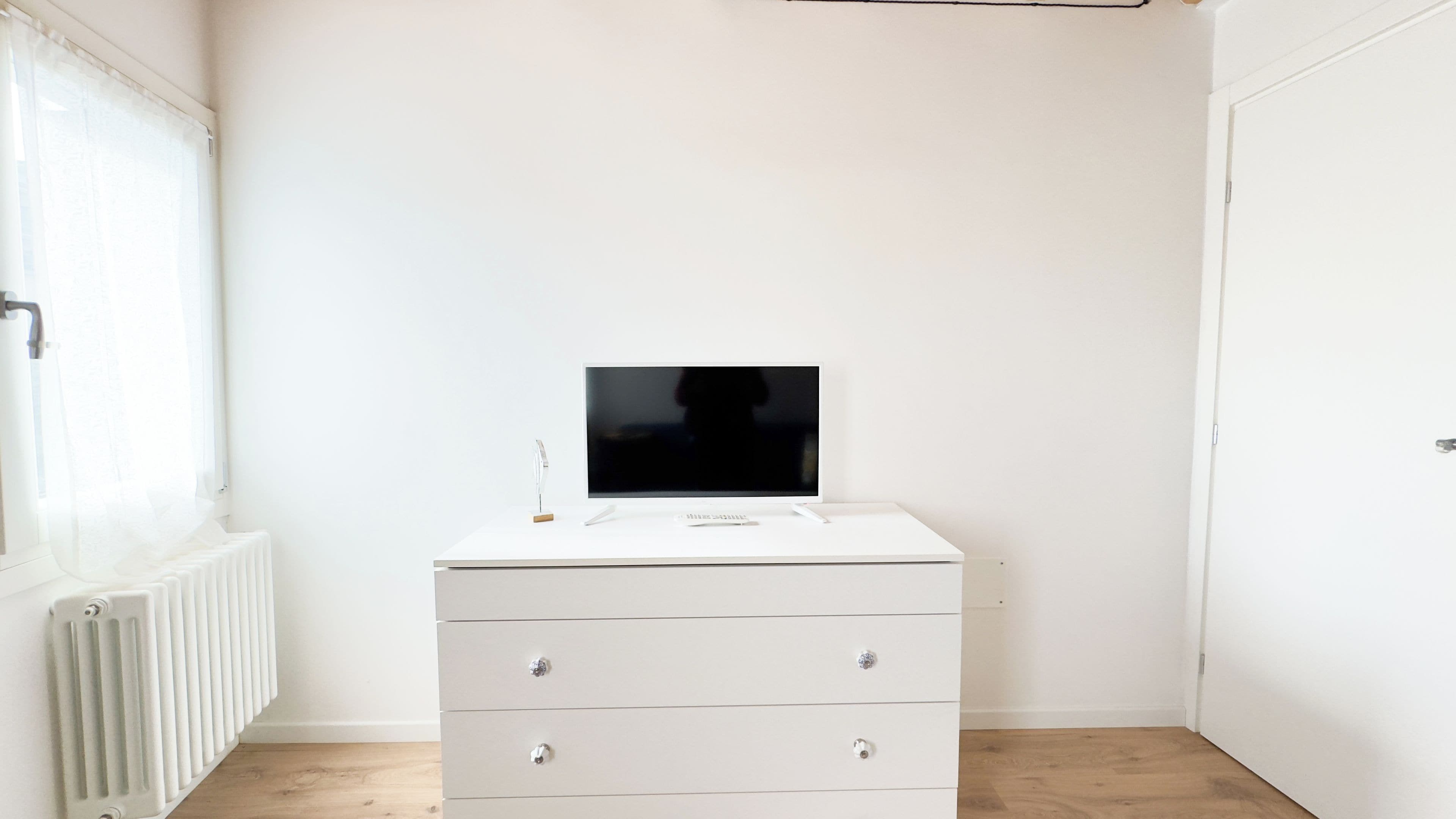 Ein weißer Kommode mit Schubladen und ein Flachbildfernseher darauf, vor einer weißen Wand mit Heizkörper darunter platziert.