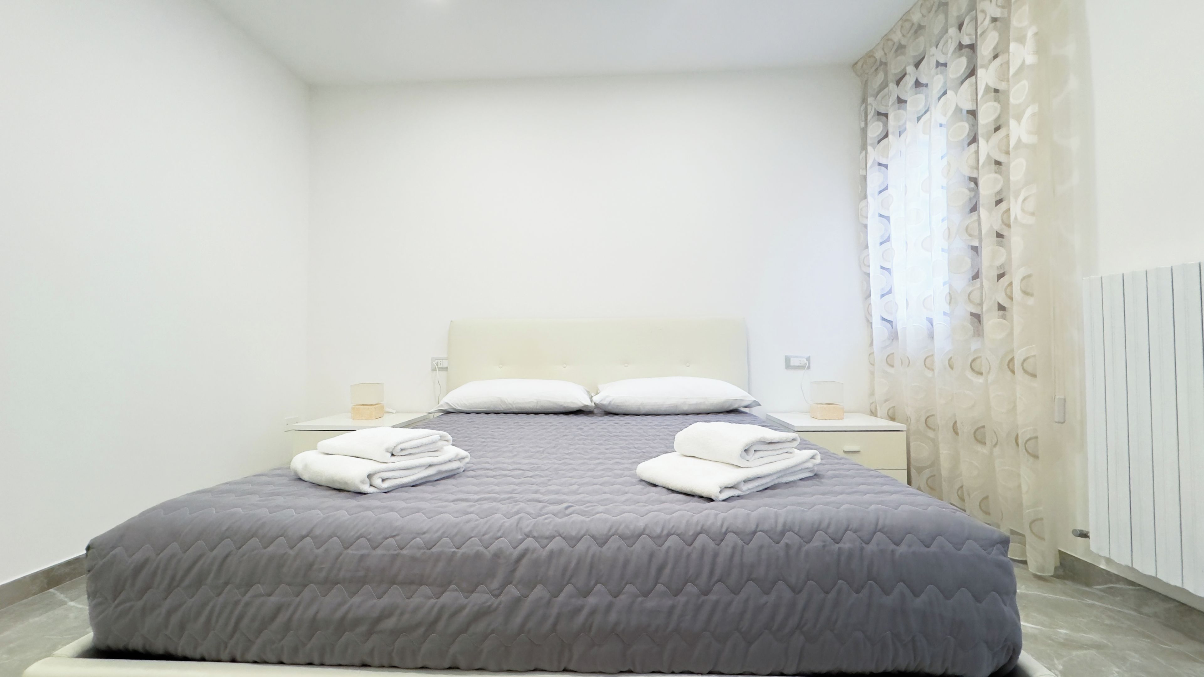 A neatly made bed with a grey quilt and two pillows, a bedside table with a glass container, and a curtain with a circular pattern.