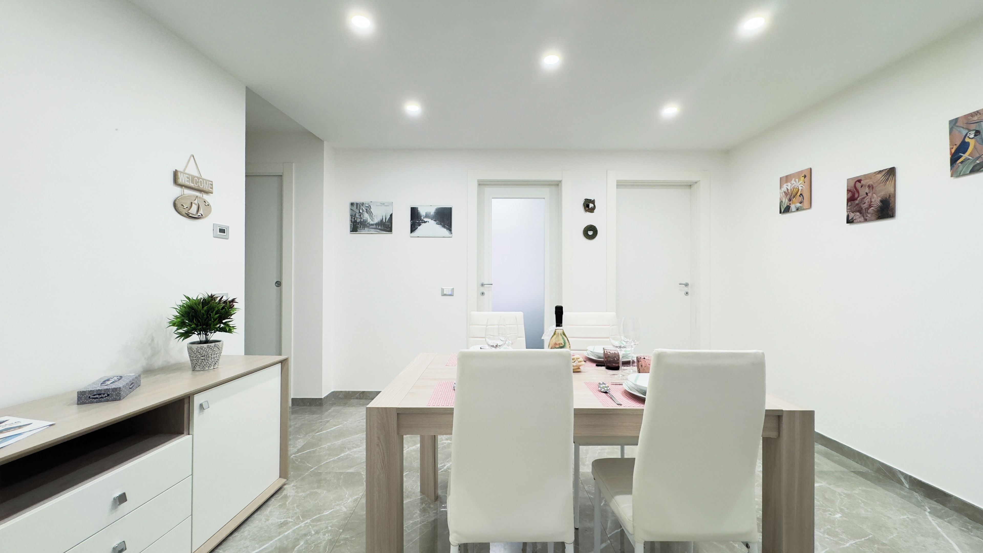 Un comedor moderno con una mesa de madera y sillas blancas, una planta en maceta y un letrero de bienvenida en la pared.