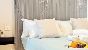 Una cama con ropa de cama blanca y azul claro, una mesita de noche con una planta y un frutero en el suelo.