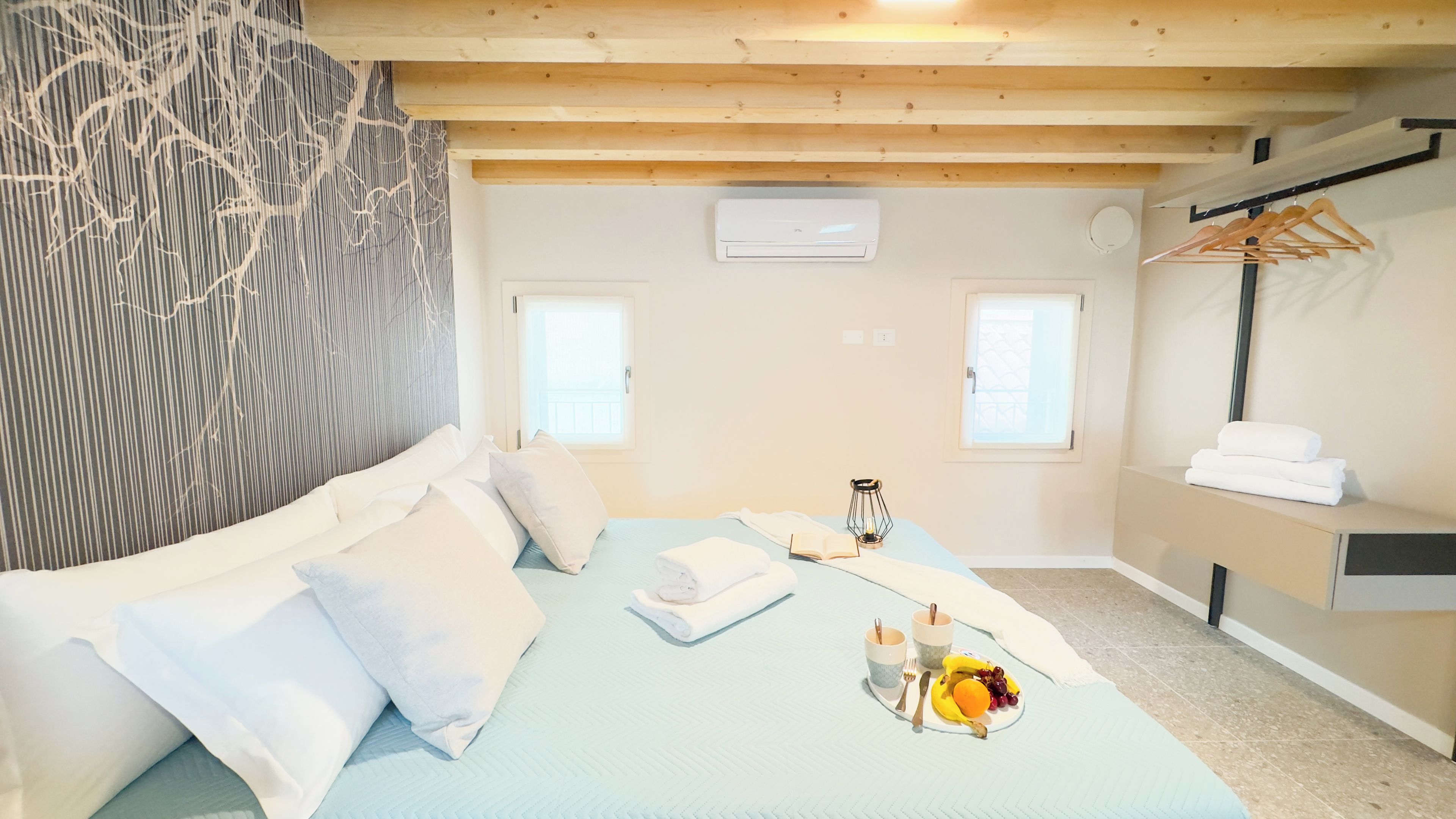 Ein Schlafzimmer mit einem Bett, Kissen, einem Nachttisch mit einer Fruitschale und einem Buch, einer Hängelampe und einem Fenster.