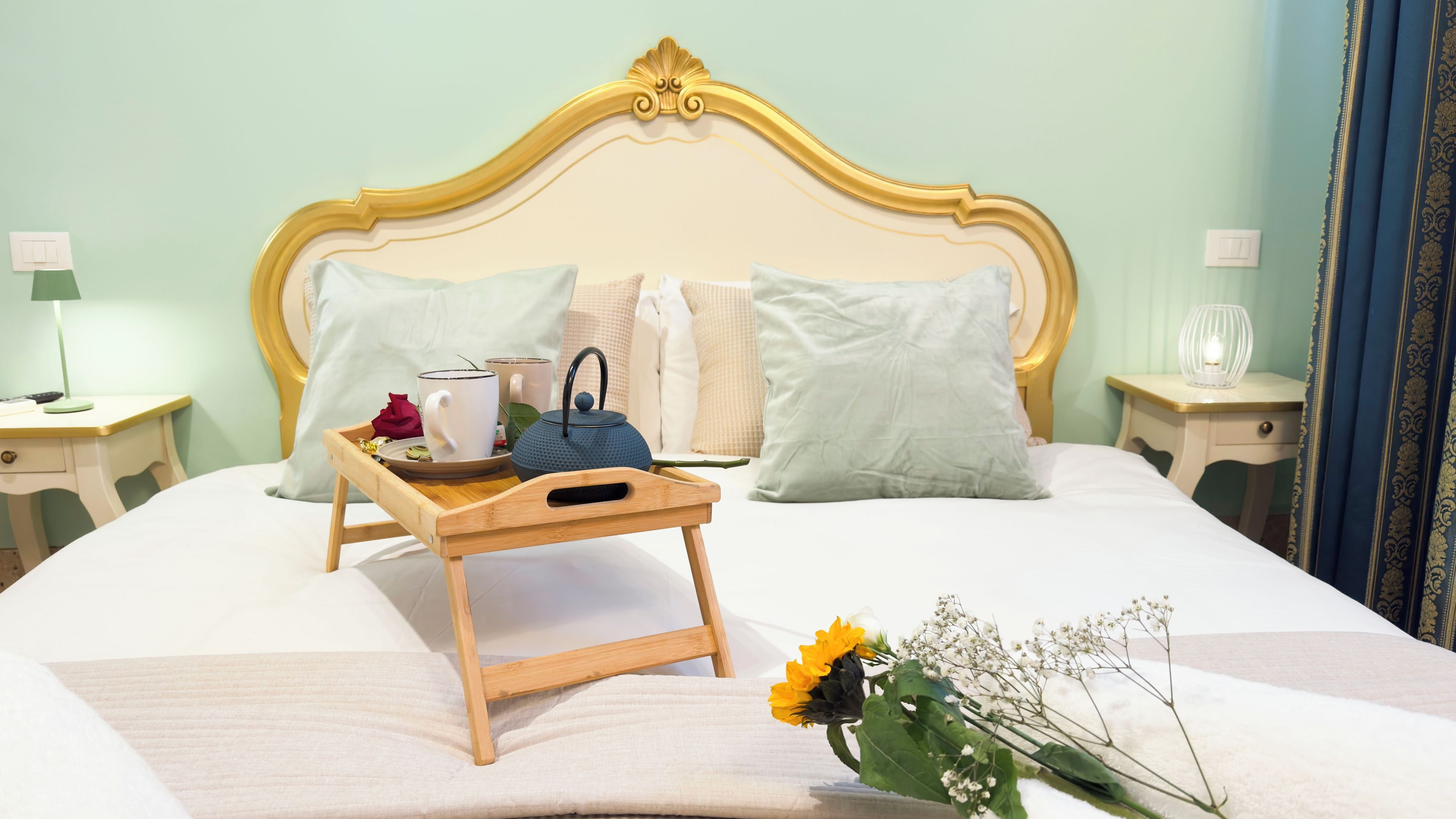 Un lit bien fait, sur lequel repose un plateau en bois contenant une théière, des tasses et une rose.