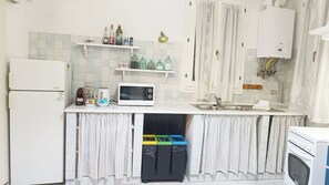 Eine kleine Küche mit Kühlschrank, Mikrowelle, Wasserkocher und Spüle. Über der Arbeitsfläche befinden sich Regale mit verschiedenen Flaschen und Behältern.