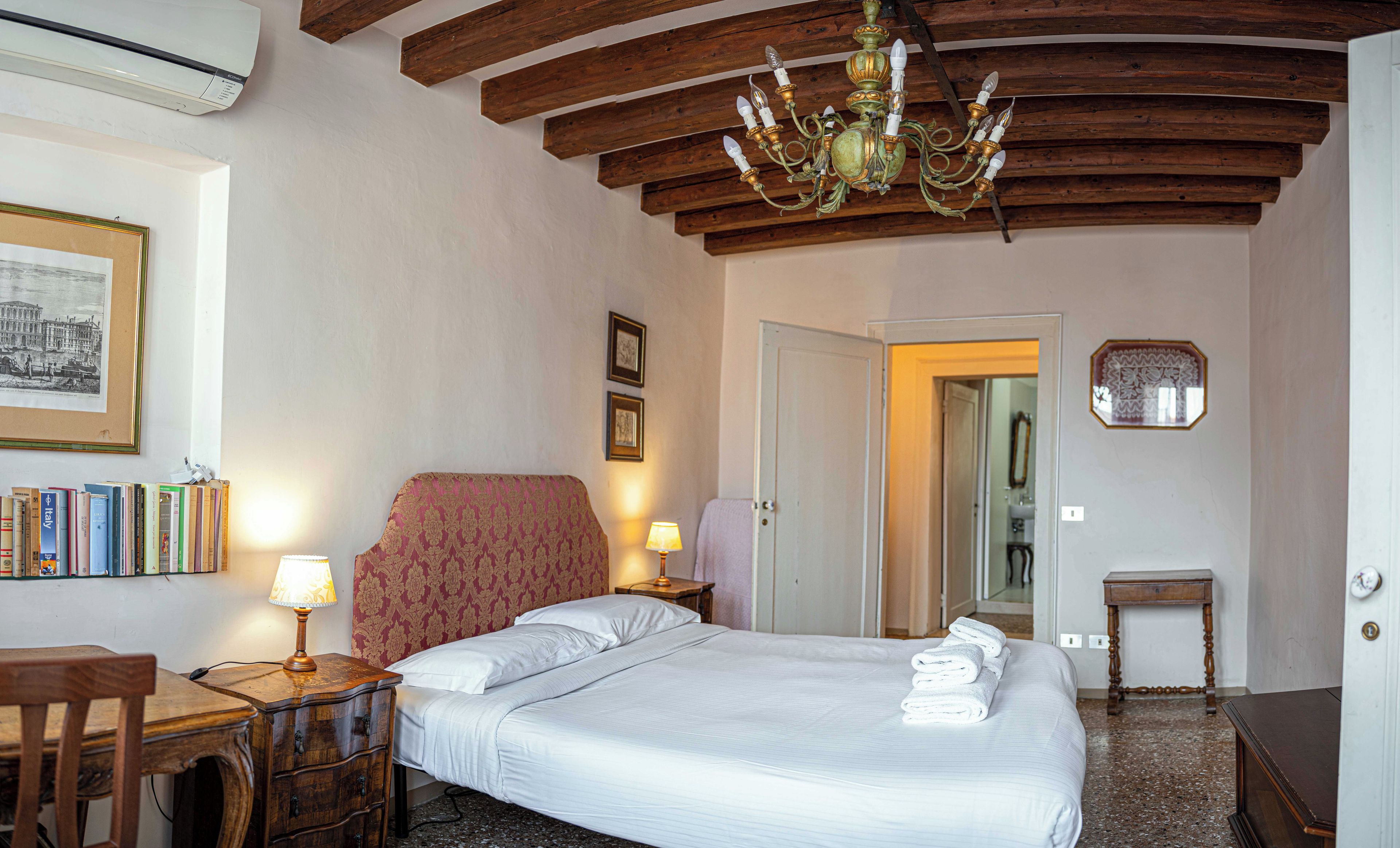 Ein Hotelzimmer mit Bett, Schreibtisch, Stuhl, Bücherregal, einer Chandelierleuchtre und einer Tür zu einem weiteren Raum.