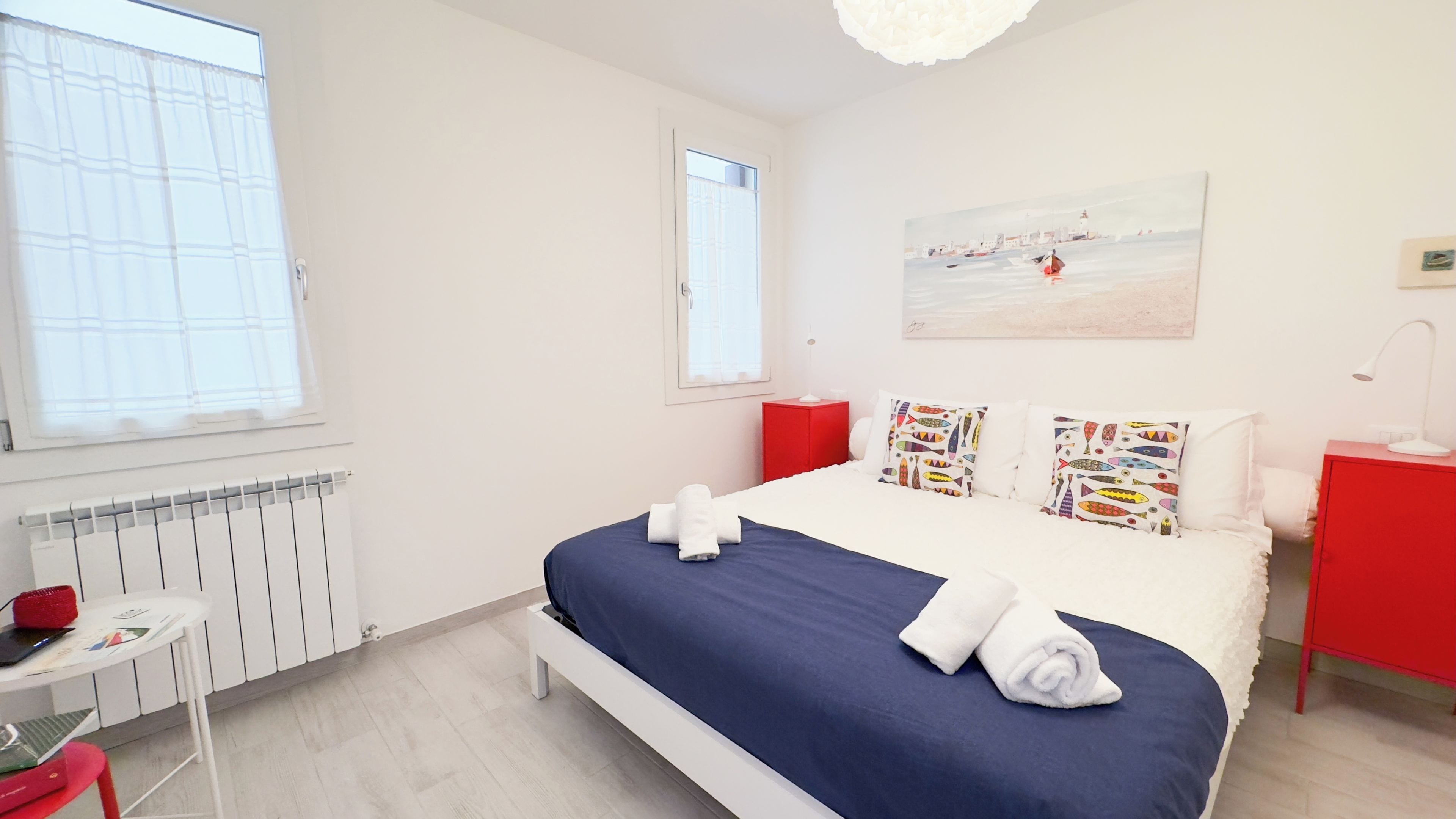 A hotel room with a bed, a red nightstand, a white desk, and a painting on the wall.