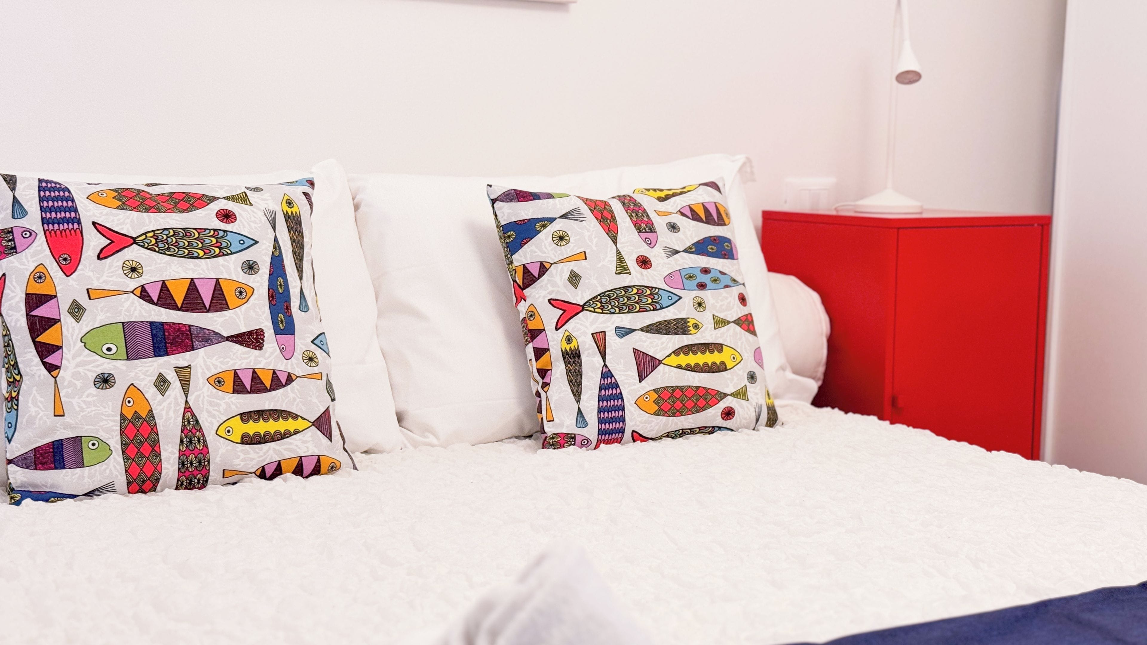 Una cama con ropa blanca y dos almohadas con motivos de peces de colores.