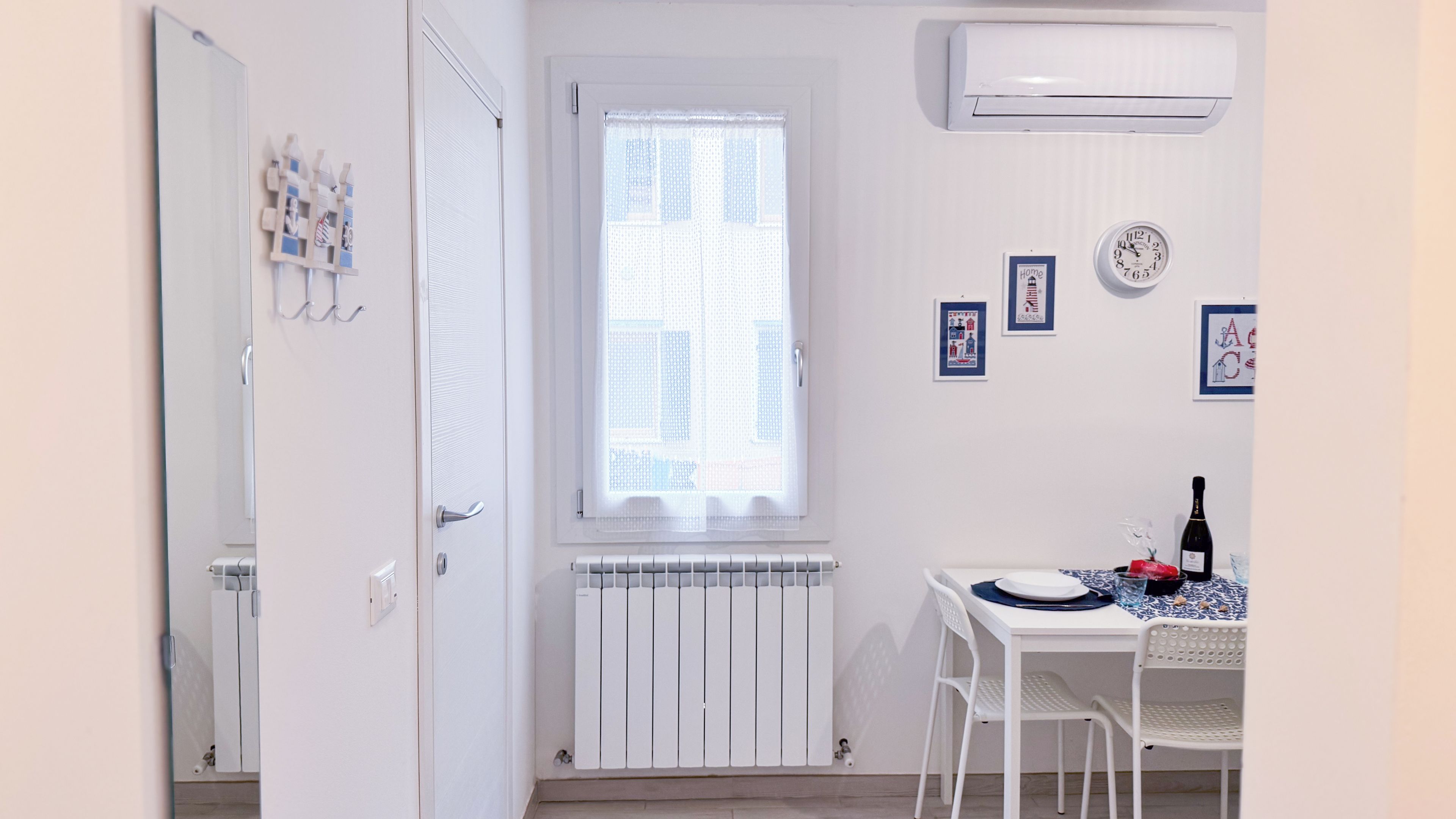 A room with a white radiator, a window with sheer curtains, a wall clock, and a table with a bottle and a plate.