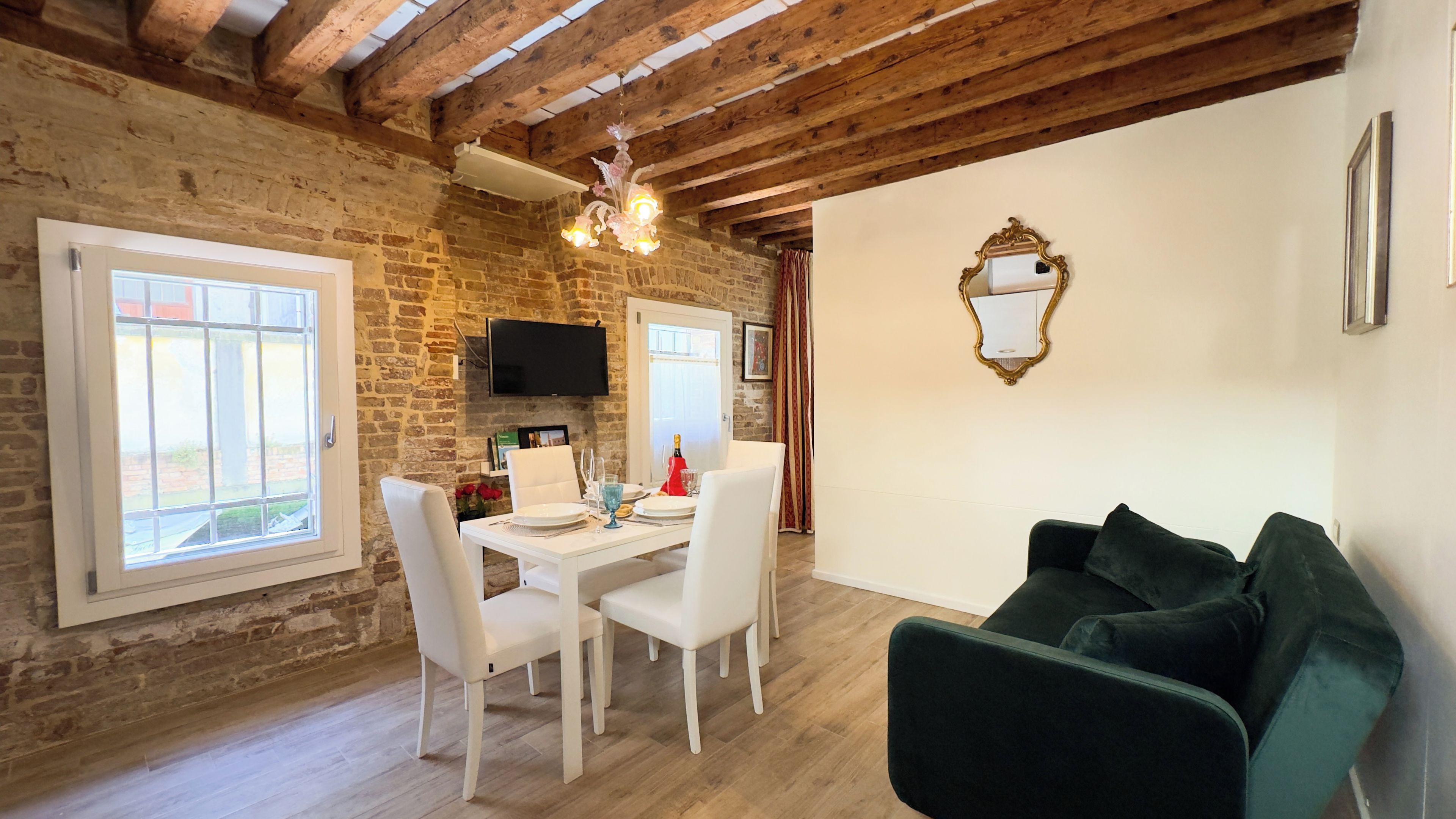 A dining area with a brick wall, wooden beams, a chandelier, a flat-screen TV, and a green sofa.